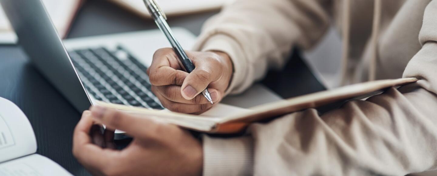 Hands writing in a notebooks with a laptop in the distance